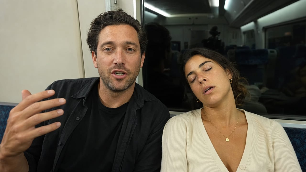 A creator talking to camera on a night train, his co-host asleep beside him
